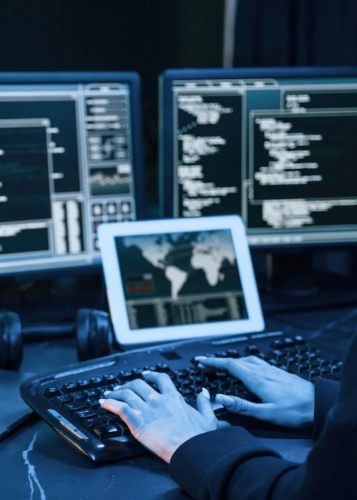 Egy személy kezei látszódnak, amint billentyűzeten gépel, előtte két számítógép monitor és egy tablet látható, mindegyiken kódok, adatok és térképpel kiegészített felhasználói felület. Sötét, éjszakai környezet. Programozás, kódolás, adatelemzés, kiberbiz