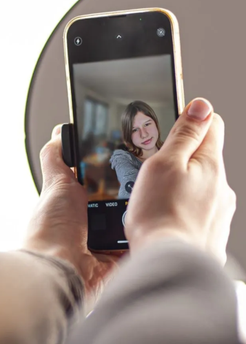 Két kéz tart egy okostelefont, amelynek kijelzőjén egy mosolygó, hosszú hajú kamasz lány szelfije látható. Szelfi, okostelefon, fotózás, portré, mobilfotó, fiatal lány, közösségi média, digitális élet, fotóapplikáció, szórakozás.