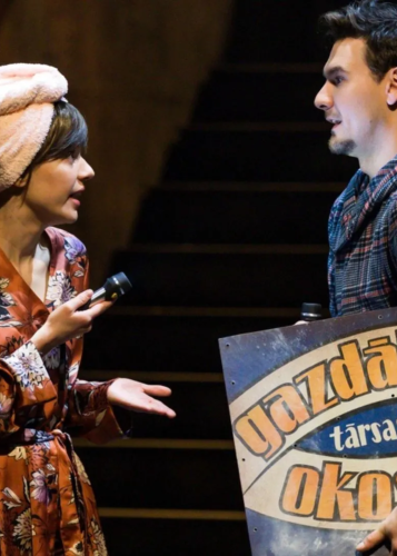 Színpadi jelenet egy előadásból: egy fürdőköntösös, turbános nő zseblámpával a kezében beszélget egy férfival, aki egy "Gazdálkodj okosan!" táblát tart. Színház, előadás, színpadi kép, szereplők, vicces jelenet, Gazdálkodj okosan, modern darab, szórakozás